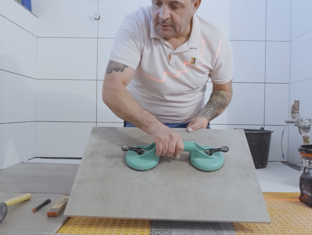 Fliesenleger legt eine große Bodenfliese in Betonoptik mit einem Saugheber präzise an ihren Platz.