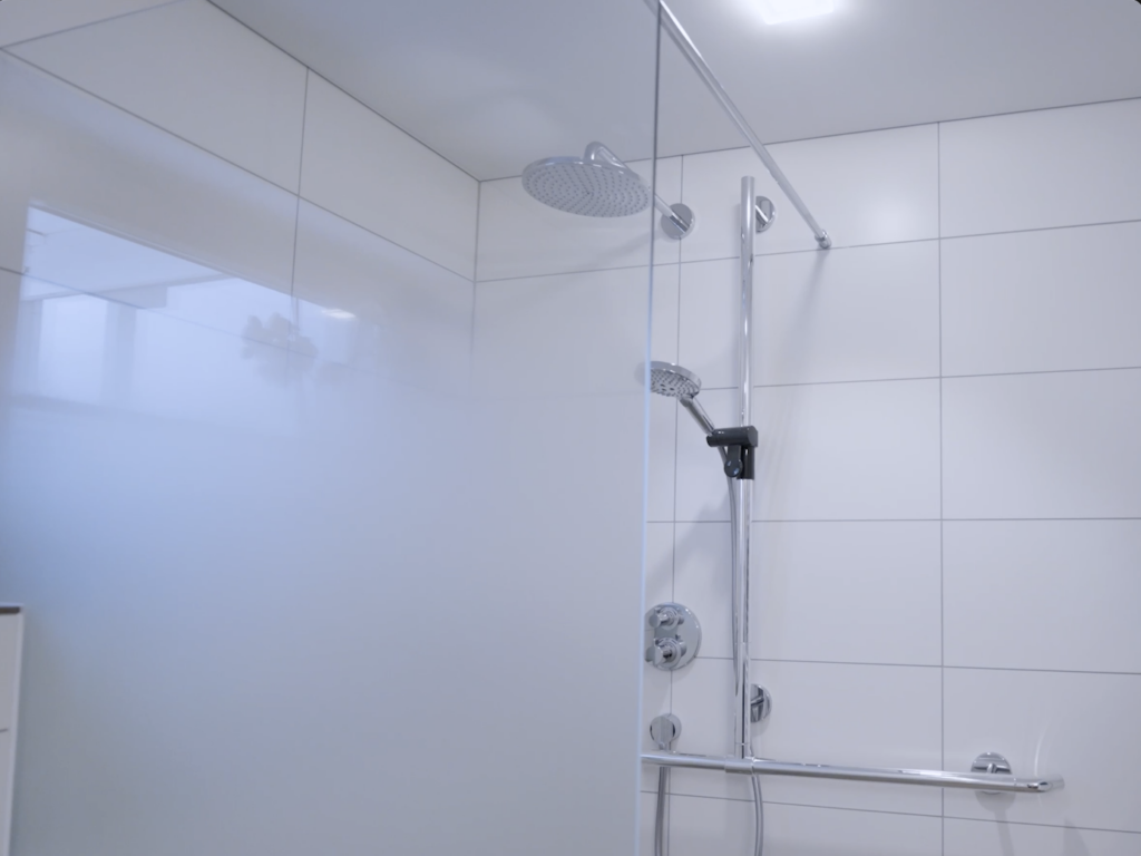 Fertige Dusche mit Glaswand, Regenbrause, Handbrause und Haltegriff vor weißen Fliesen.