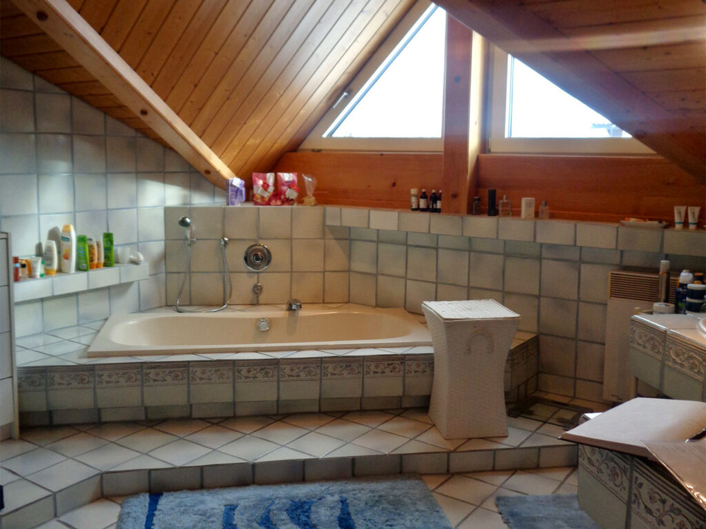 Vorher: Dachgeschoss-Bad mit Holzdecke, kleinen weißen Fliesen mit Blumenbordüre und einer Badewanne auf einem gefliesten Podest mit Stufe.