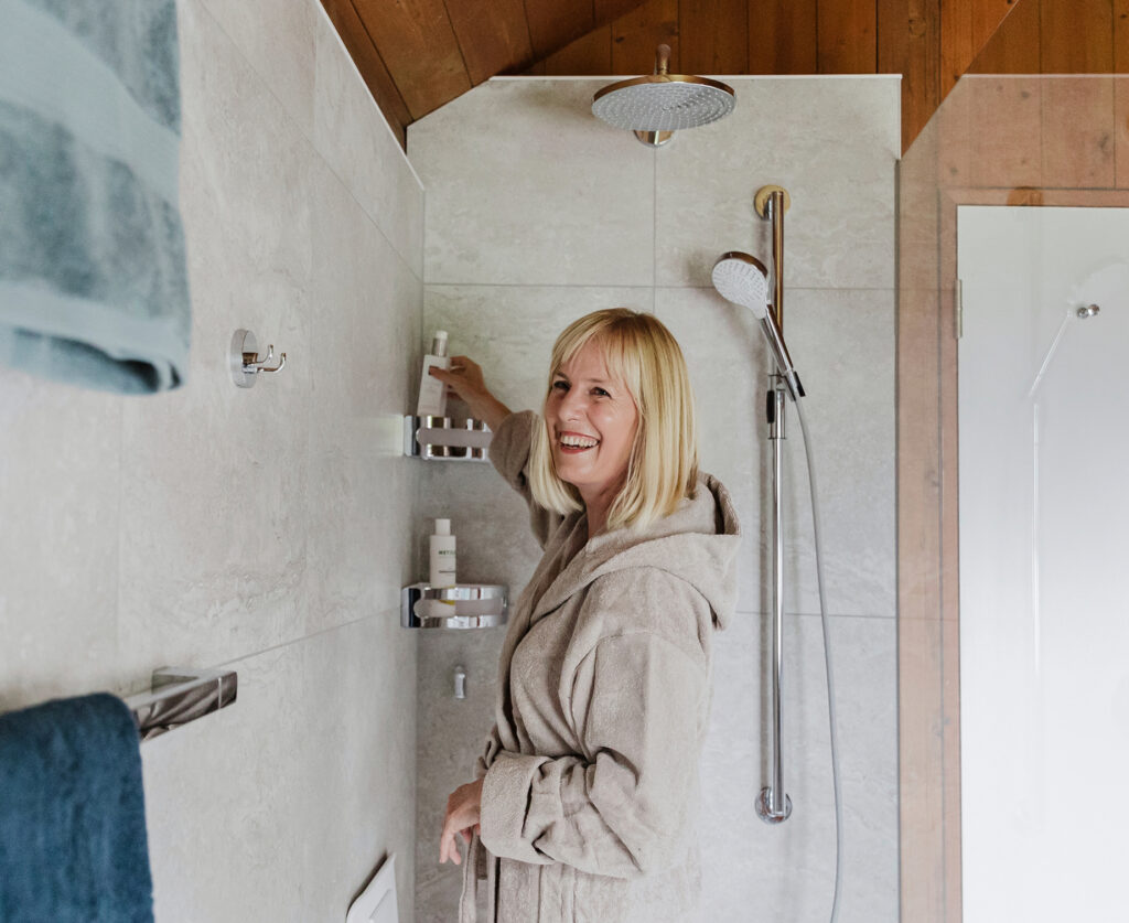 Lächelnde Frau im Bademantel in einer neuen Dusche mit Regenbrause, Handbrause und Ablagen unter einer Dachschräge aus Holz.