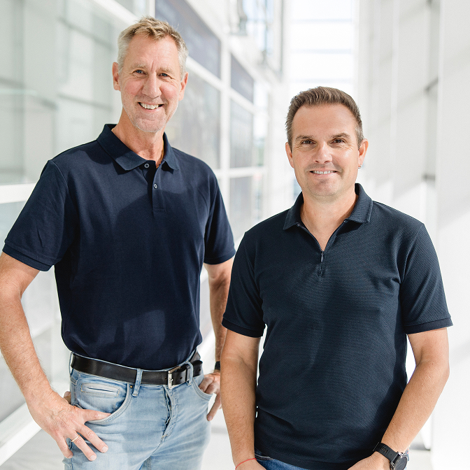 Die beiden Vollbad-Ansprechpartner stehen lächelnd nebeneinander in dunklen Poloshirts vor einer hellen Glasfassade.
