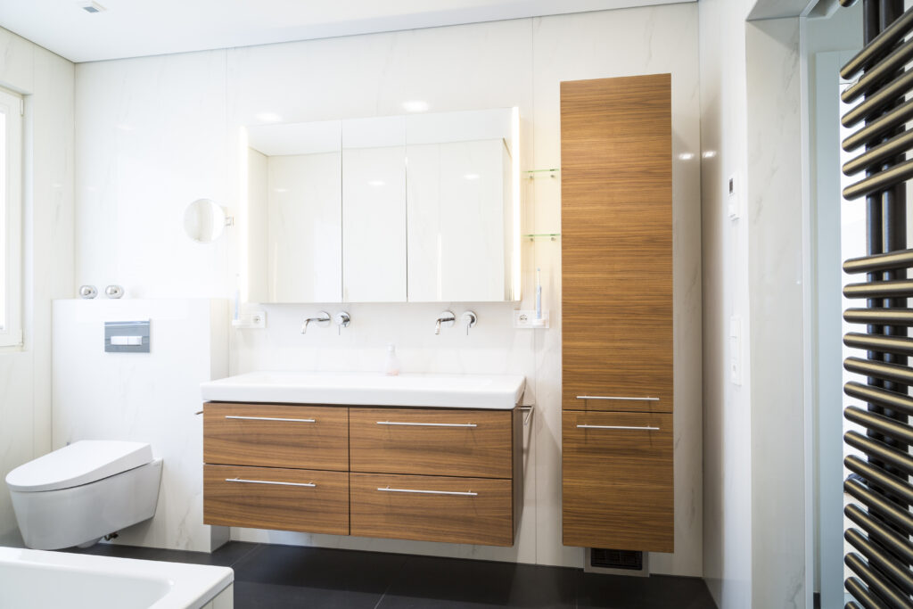 Bad in Klaus mit Doppelwaschbecken auf Nussholz-Unterschrank, beleuchtetem Spiegelschrank, Hochschrank und Dusch-WC vor hellen Fliesen.