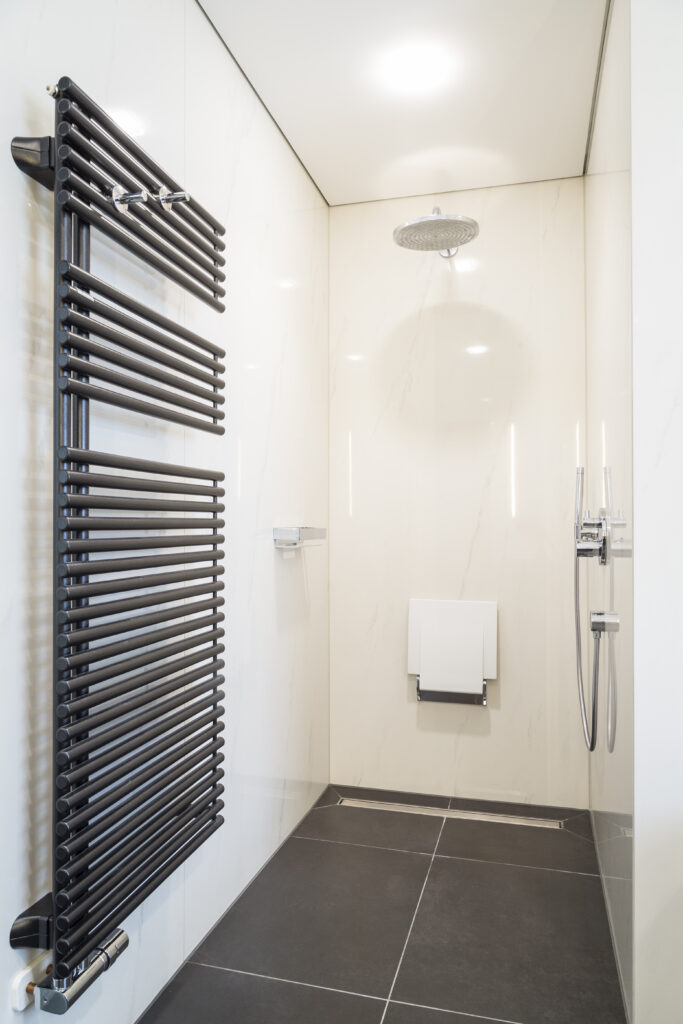Bodengleiche Dusche mit Rinne, Regenbrause und Klappsitz, davor ein anthrazitfarbener Handtuchheizkörper.