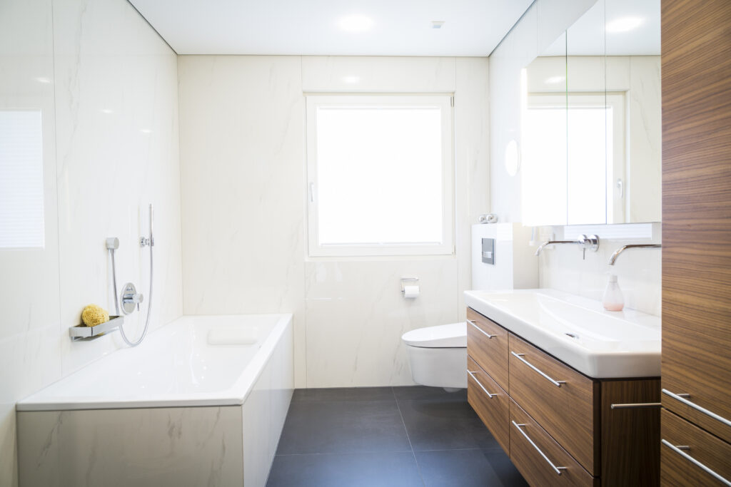 Helles Bad mit eingefliester Badewanne, Hänge-WC unter dem Fenster und Waschtisch mit Nussholz-Schubladen auf dunklem Boden.