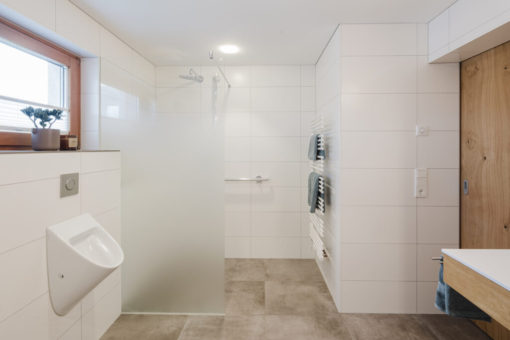 Barrierefreies Bad in Rankweil: bodengleiche Dusche mit satinierter Glaswand und Haltegriff, daneben Urinal und weißer Handtuchheizkörper.