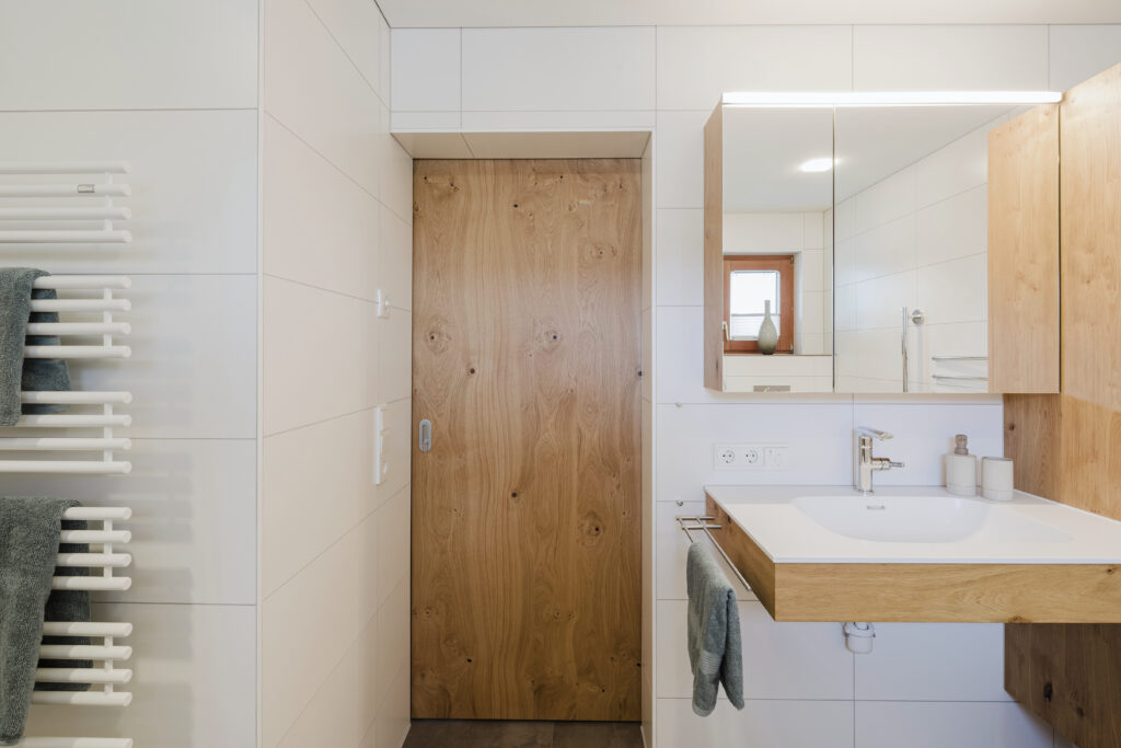 Unterfahrbarer Waschtisch mit Eichenholz-Unterbau und Spiegelschrank, daneben eine Schiebetür aus Eiche.