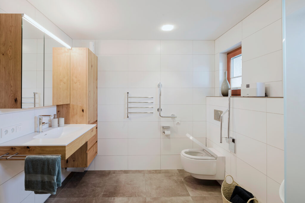Helles Bad mit Waschtisch und Hochschrank aus Eiche, gegenüber ein Hänge-WC mit Stützklappgriff und Haltegriff an der Wand.