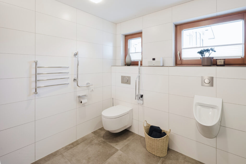WC-Bereich mit Dusch-WC, hochklappbarem Stützgriff, Winkelgriff und Urinal unter zwei Holzfenstern.
