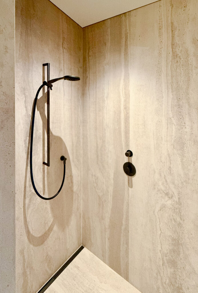 Bodengleiche Dusche mit schwarzer Handbrause an der Stange und schwarzem Thermostat vor warm beleuchteten Wänden in Travertinoptik.