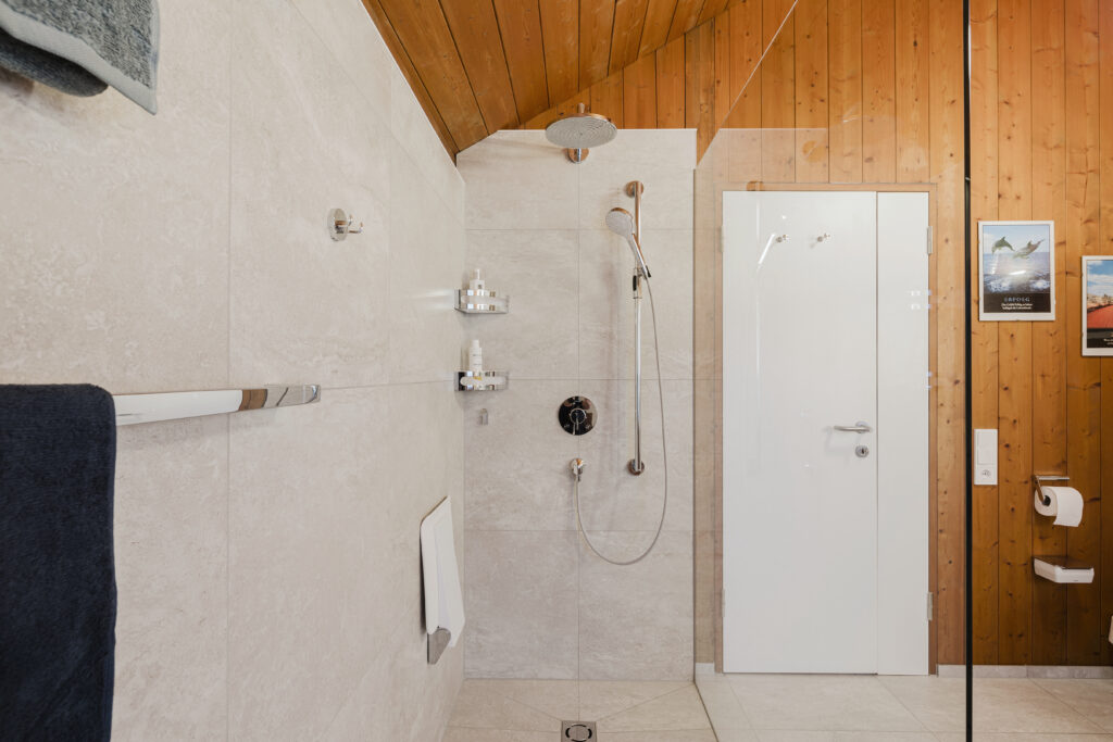 Blick in die bodengleiche Dusche mit Regenbrause, Handbrause an der Haltestange, Klappsitz und Ablagen unter der Holzdecke.