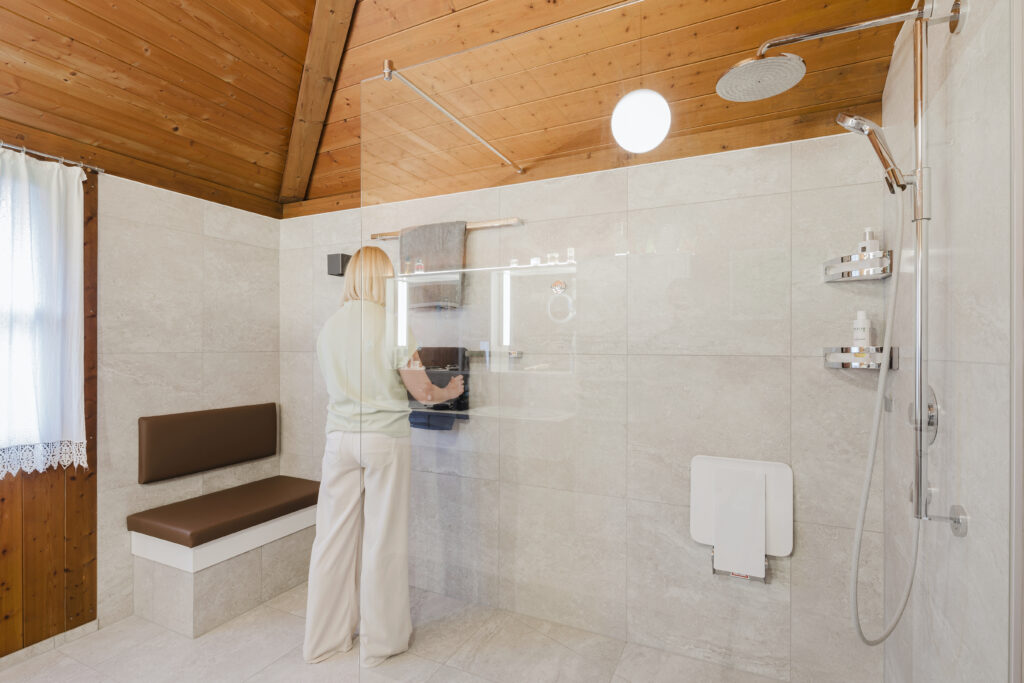 Frau hängt ein Handtuch hinter der Glaswand der Dusche auf, daneben eine gepolsterte Sitzbank in hellen Fliesen unter der Holzdecke.