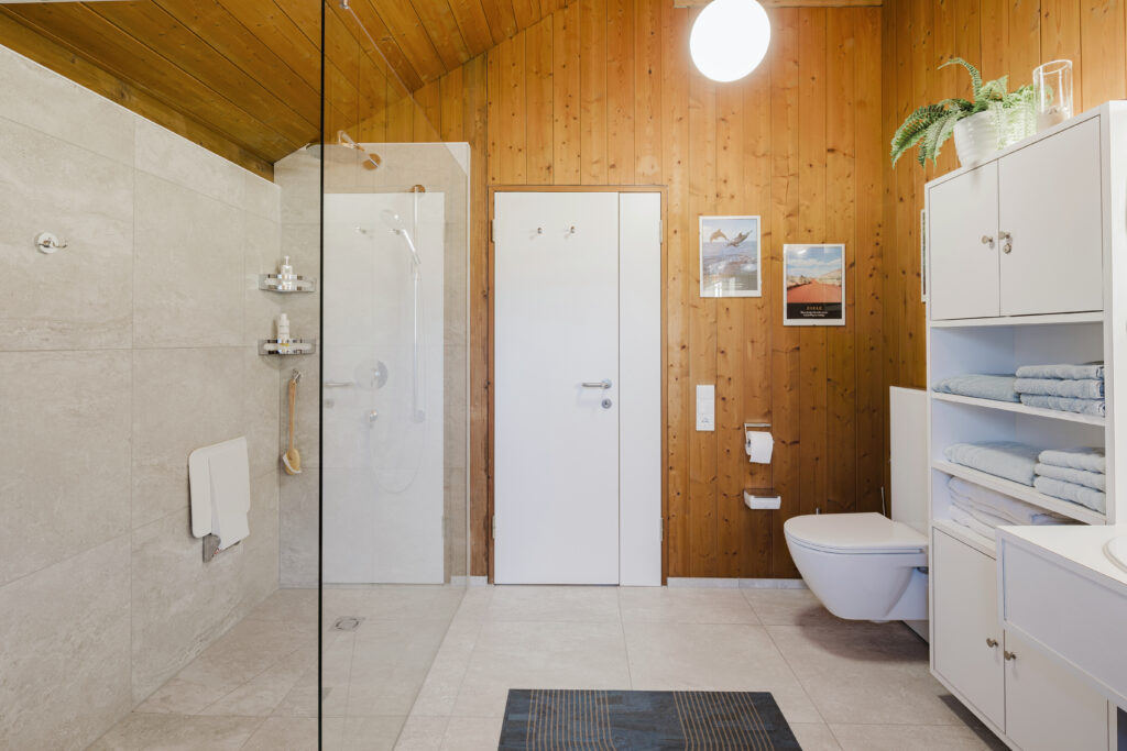 Teilsaniertes Bad in Hohenems: bodengleiche Dusche mit Glaswand und Klappsitz, helle Fliesen und Hänge-WC, die Holzwände blieben erhalten.
