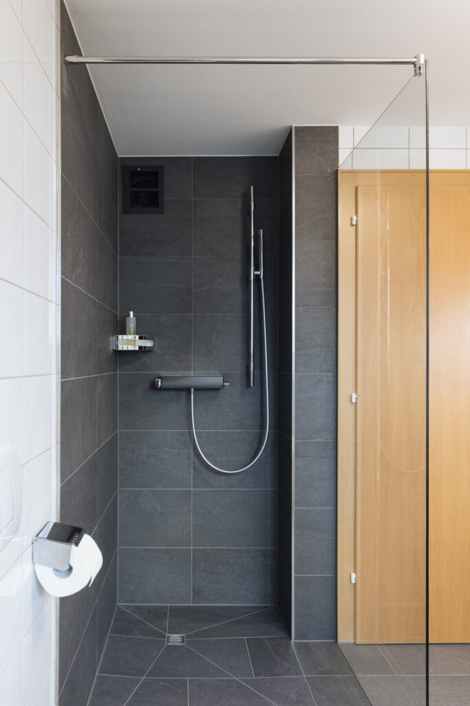 Frontalansicht der Dusche mit Glaswand, Thermostat, Handbrause an der Stange und Eckablage, Bodenablauf in der Mitte.