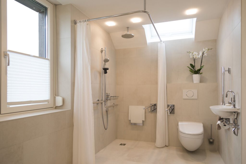 Gesamtansicht von Dusch- und WC-Bereich mit Regenbrause, Klappsitz, Stützgriffen und kleinem Handwaschbecken, alles stufenlos erreichbar.
