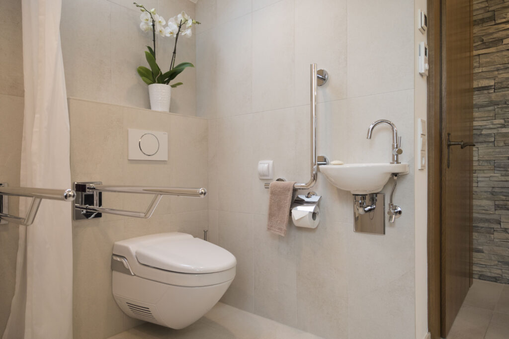 Dusch-WC mit hochklappbarem Stützgriff, daneben Handwaschbecken mit Winkelgriff und Papierhalter in Griffhöhe.