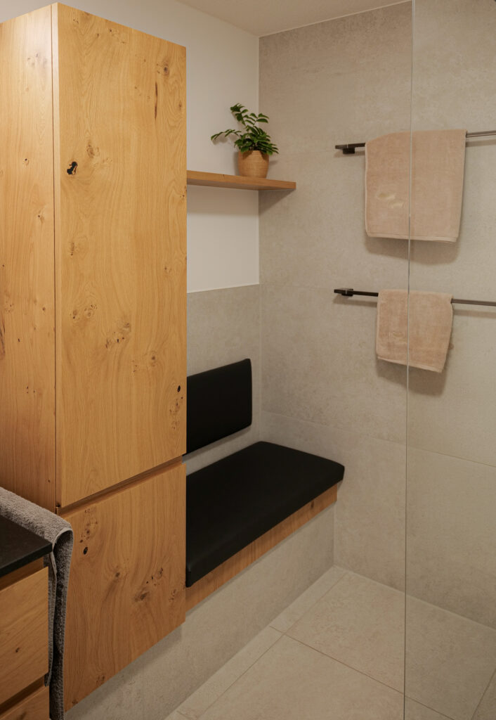 Bad in Feldkirch mit Hochschrank aus Asteiche, gepolsterter Sitzbank und Wandregal neben der bodengleichen Dusche mit Glaswand.