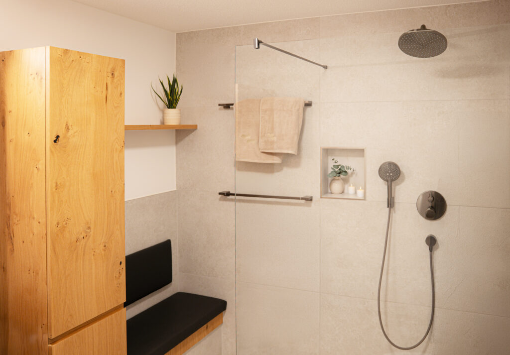 Walk-in-Dusche mit Regenbrause, Handbrause und Armaturen in dunklem Metall, dazu eine Wandnische mit Kerzen und Handtuchstangen.