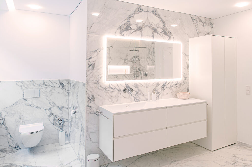 Komplett saniertes Bad in Bregenz: weißer Doppelwaschtisch mit Wandarmatur vor Marmorwand, hinterleuchteter Spiegel und separater WC-Bereich.