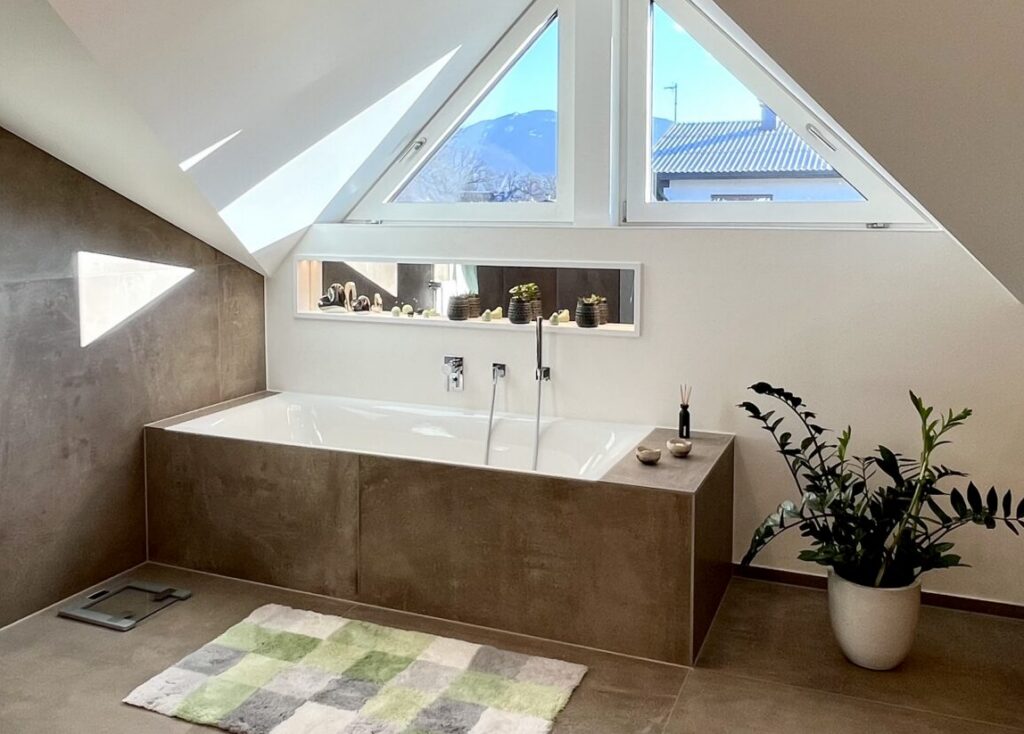 Weitwinkelblick auf eingeflieste Badewanne unter dreieckigen Giebelfenstern, links die Glasdusche, rechts Eichenmöbel, Boden in Betonoptik.