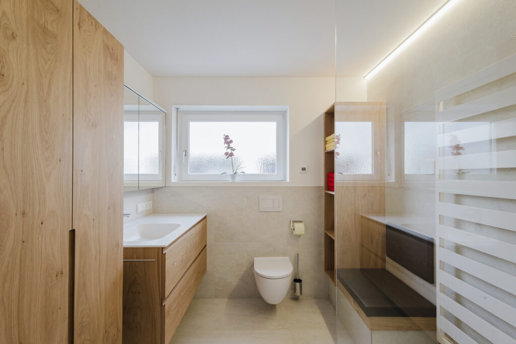 Helles Bad in Rankweil mit Hochschrank und Waschtisch aus Eiche, Hänge-WC unter dem Fenster und gepolsterter Sitzbank mit offenem Regal.