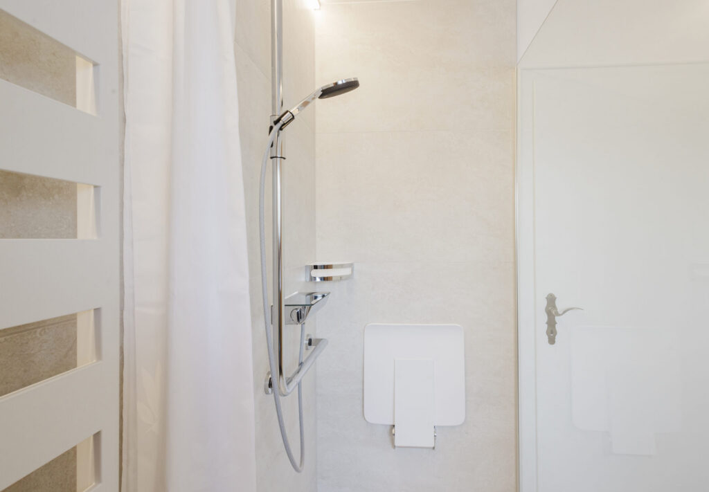 Nahaufnahme der Dusche mit Thermostat, Ablage, Haltegriff und weißem Klappsitz vor hellen Fliesen.