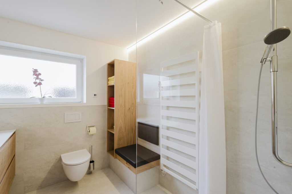 Hänge-WC unter dem Fenster, daneben offenes Eichenregal mit Handtüchern, gepolsterte Sitzbank und die Dusche mit Glaswand.