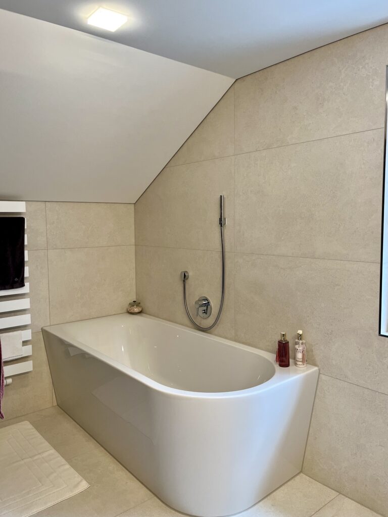 Weiße Vorwand-Badewanne mit abgerundetem Ende unter der Dachschräge, umgeben von großen beigen Fliesen in Steinoptik.