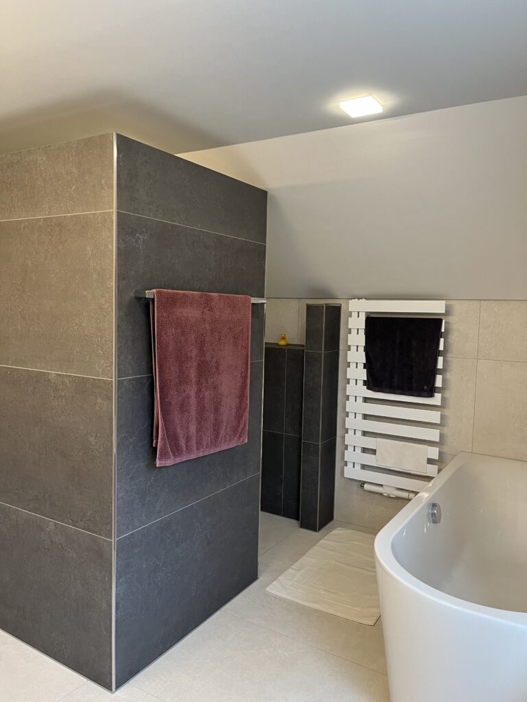 Dunkel geflieste Duschtrennwand mit Handtuchhalter, daneben ein weißer Designheizkörper und die Badewanne.