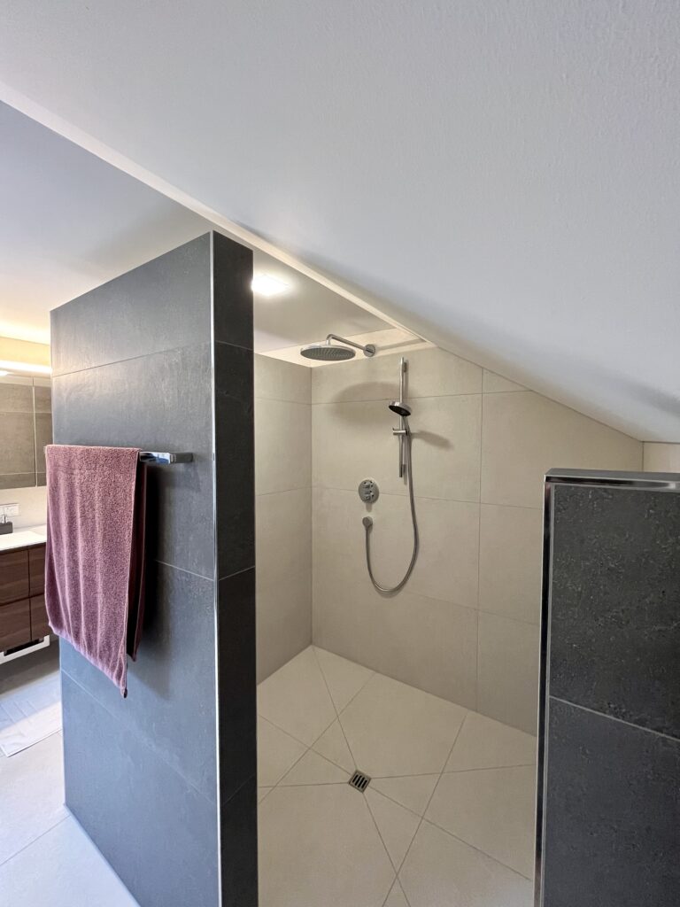 Offene, bodengleiche Walk-in-Dusche unter der Dachschräge mit Regenbrause, Handbrause und dunklen Trennwänden.