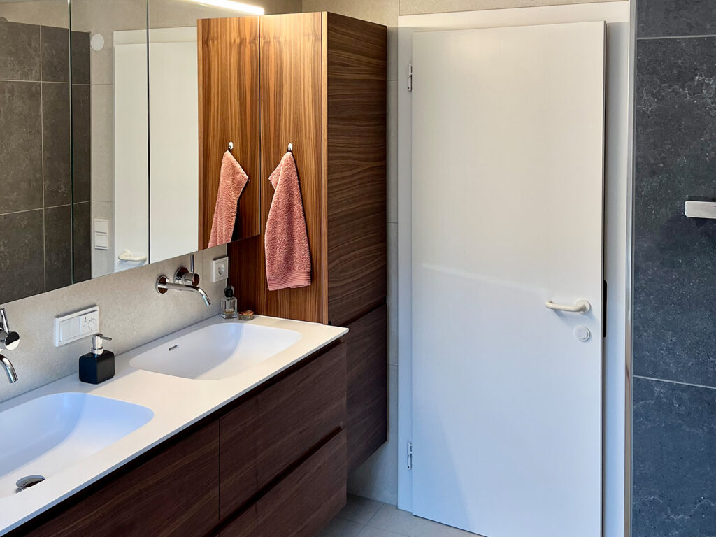 Nachher: Doppelwaschtisch mit Unterschrank und Hochschrank in Nussholz, großer Spiegelschrank und weiße Tür, Fliesen in Beige und Anthrazit.