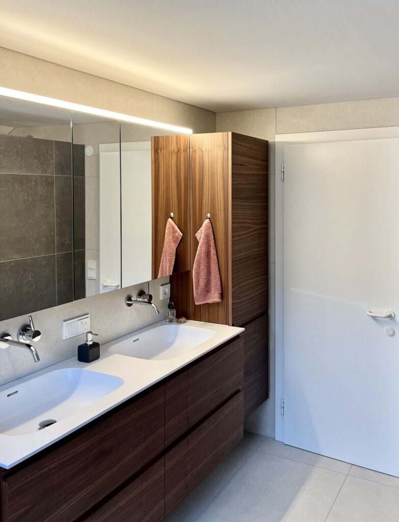 Heller Waschbereich mit zwei weißen Becken, Wandarmaturen, dunklen Nussholzfronten und langem Spiegelschrank mit Lichtleiste.