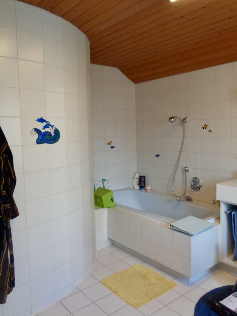 Vorher: Bad in Feldkirch mit Holzdecke, kleinen weißen Fliesen mit Fisch-Motiven, runder Duschwand und eingefliester Badewanne.