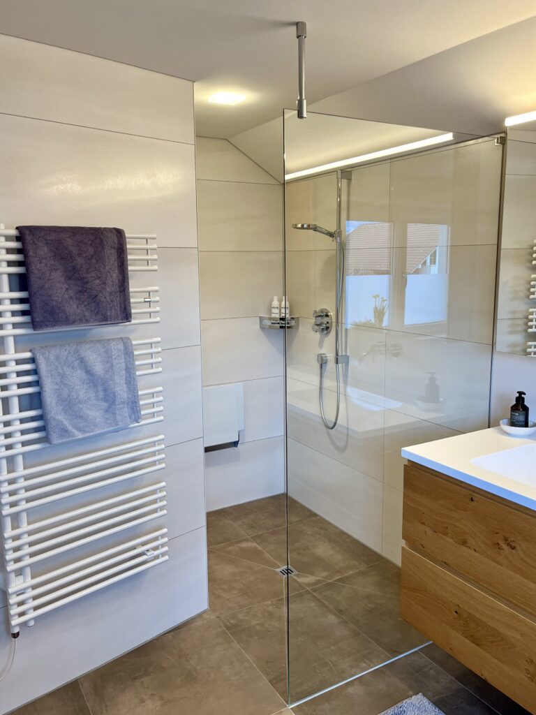 Bodengleiche Dusche mit Glaswand, Handbrause an der Stange und Klappsitz, daneben ein weißer Handtuchheizkörper.