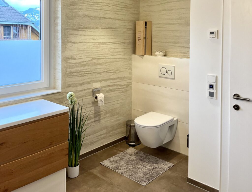 Hänge-WC vor heller Vorwand mit Ablage, daneben eine Wand mit Strukturputz und ein Fenster mit Bergblick.