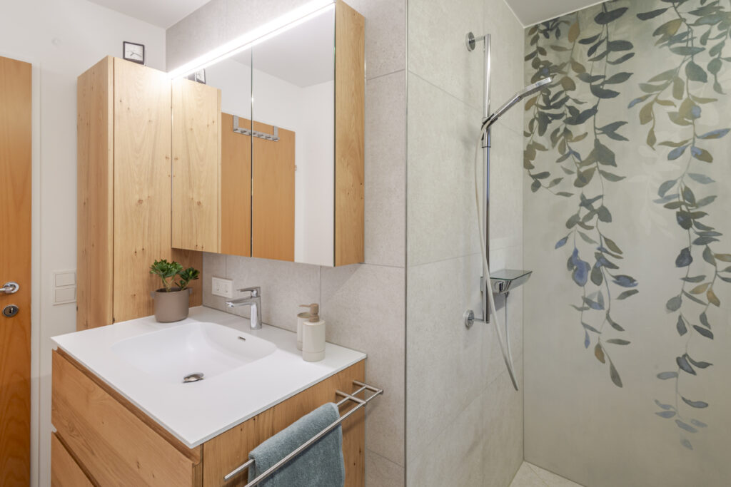 Bad in Hard mit Waschtisch und Hochschrank aus Eiche, beleuchtetem Spiegelschrank und Dusche mit floral gemusterter Rückwand.