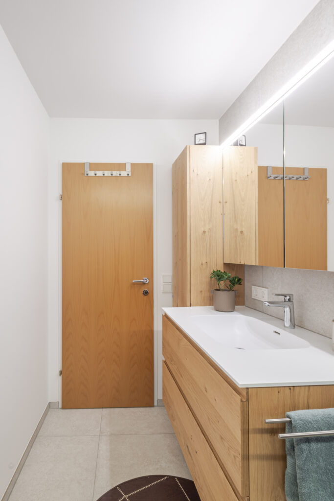 Blick auf Waschtisch und Hochschrank aus Eiche neben der Zimmertür, darüber der Spiegelschrank mit Lichtleiste.