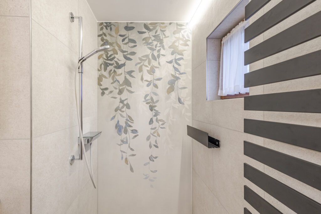 Dusche mit Handbrause, Glasablage und einer Rückwand mit hängendem Blättermotiv, daneben der schwarze Heizkörper.