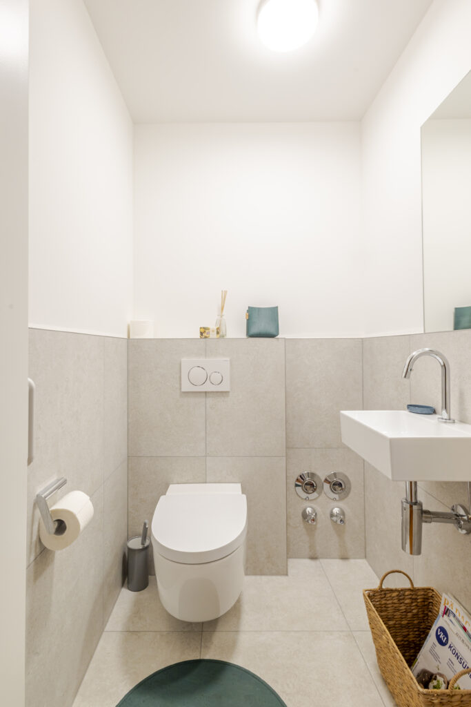 Separates WC mit Hänge-WC, kleinem Waschbecken, Spiegel und hellen Fliesen in Steinoptik.