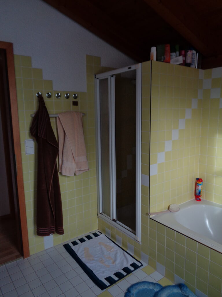 Vorher: enge Duschkabine mit Falttür und Stufe, umgeben von kleinen gelben Fliesen, daneben die eingeflieste Badewanne.