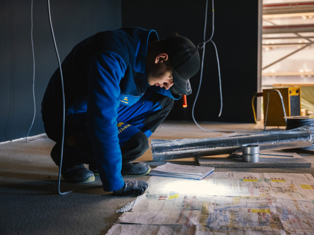 Elektriker vom Dorfelektriker kniet auf der Baustelle und prüft mit einer Taschenlampe den Installationsplan am Boden.
