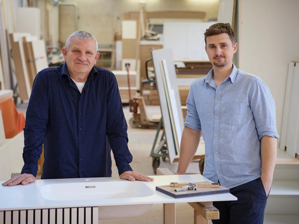 Zwei Tischler der Tischlerei Schwab stehen in ihrer Werkstatt an einem weißen Waschtisch mit Holzlamellen-Unterbau.